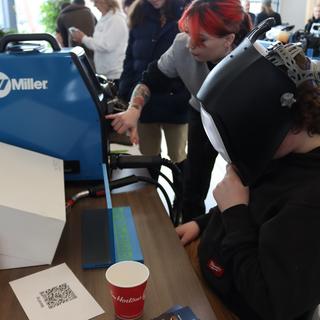 A person wearing a welding helmet is sitting at a desk trying a welding simulator machine