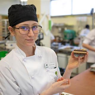 A person wearing a chef hat and chef jacket is holding up a Nanaimo bar dessert.