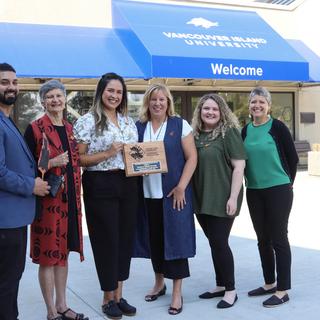 A group stands in front of the welcome centre holding a plaque
