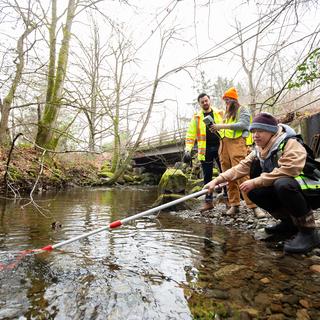 Three people next to a stream, one holds a sampling pole while the others look on
