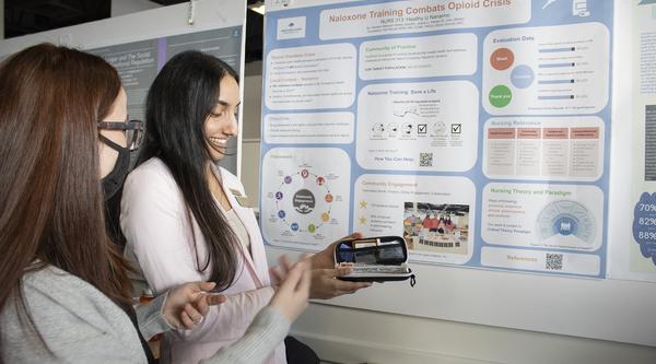 Two students discuss a poster behind them, one holds a naloxone kit