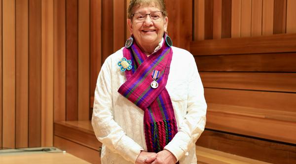 Métis Elder-in-Residence Stella Johnson standing in a cedar panelled room wearing a red and purple tartanMétis scarf with a King Charles III Coronation Medal