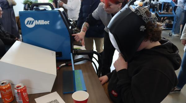 A person wearing a welding helmet is sitting at a desk trying a welding simulator machine