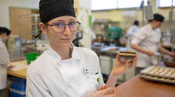 A person wearing a chef hat and chef jacket is holding up a Nanaimo bar dessert.