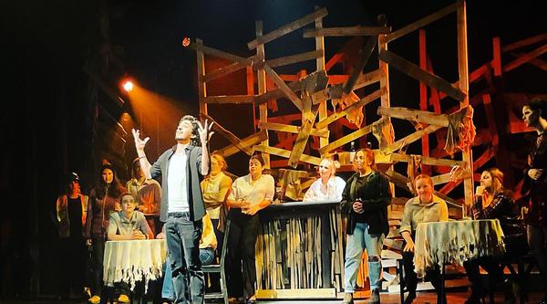 An actor at centre stage has their hands thrown up in the air while they look off in the distance. Numerous cast mates stand behind them in various poses.