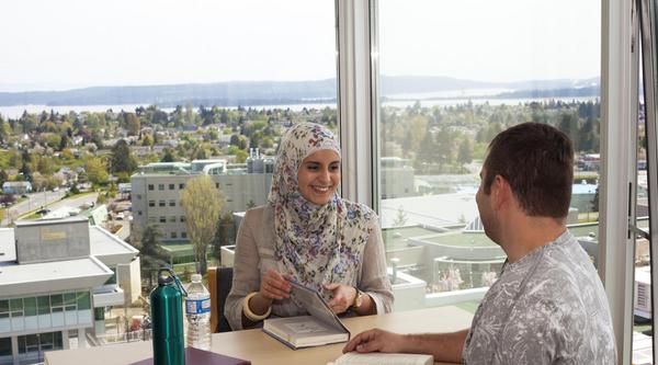 man and woman smiling while sitting across from one another at a table by a window. textbooks are on the desk in front of them