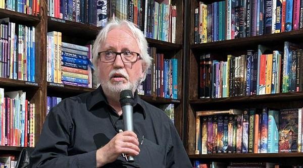 Perry Bulwer speaks into a microphone with a shelf of books behind him