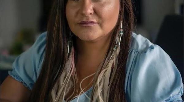 Brandi is wearing a blue shirt and sits with her arms on a table in front of her