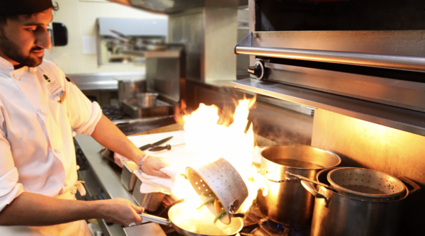 Culinary Student pouring vegetables into hot pan to sear them, creating short burst of flames in the pan