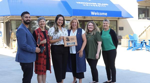 A group stands in front of the welcome centre holding a plaque