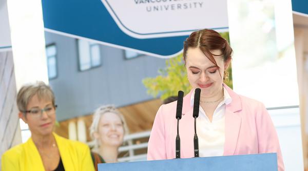 VIU student Mallory Woods stands at a podium wearing a pink blazer. VIU banners stand behind them.