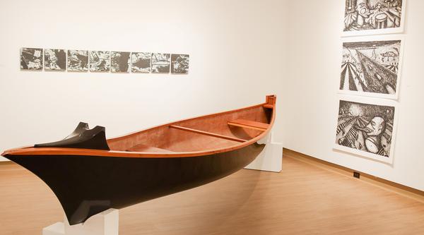 A canoe sits on a stand with drawings hanging on the gallery wall's behind it.