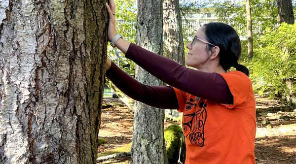 A woman wearing an orange t-shirt for Truth and Reconciliation Day putting her hands on a large tree in an act of gratitude/
