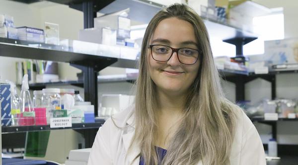 Kimberley Barrett wears a white lab coat and stands in a research lab.