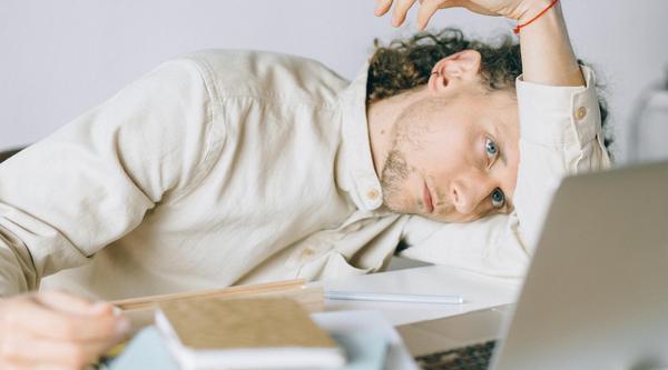 Student leaning on his arm staring dejectedly at a laptop screen with a pile of books beside him
