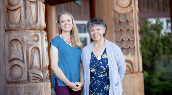 VIU Psychedelic-assisted Therapy Program Chair Shannon Dames with VIU Elder-in-Residence C-tasi:a Geraldine Manson standing outside in front of totem poles and smiling at the camera