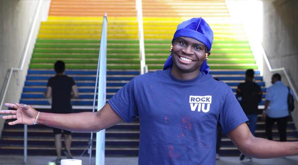 Black student holds arms out in welcome standing in front of the rainbow stairs