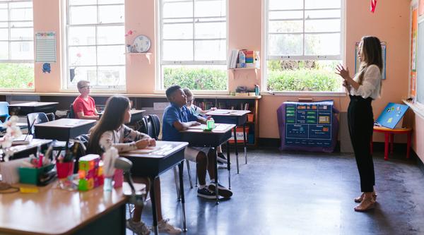 Students sit at desks in a class listening to a teacher