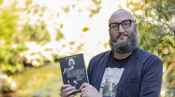Dr. Theo Finigan, a VIU English Professor, holds up the book Atomic Blonde while standing in front of the VIU koi pond.