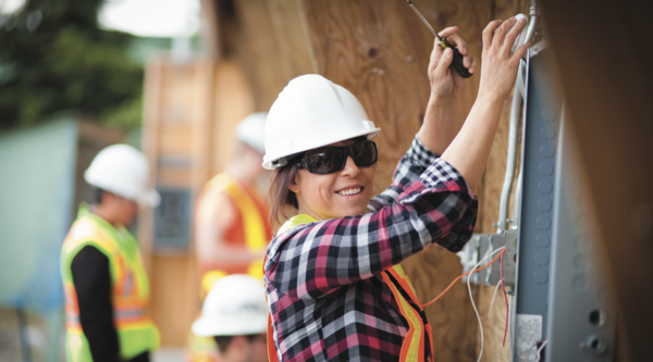 Female electrician wearing. white hard hat a safety vest and sunglasses working on the side of a building and smiling at the camera.
