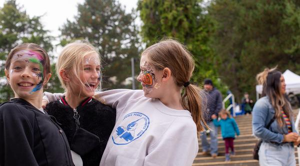 Three young girls wearing face paint hug, with a crowd on the stairs behind them