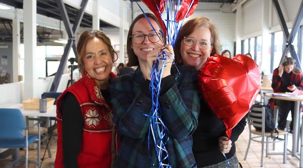 Three women smile, one holding a bunch of balloons, another holding a heart balloon near her face