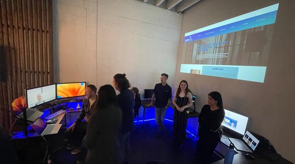 Students gather around three screens on a desk with a projection of an image on the wall behind them.