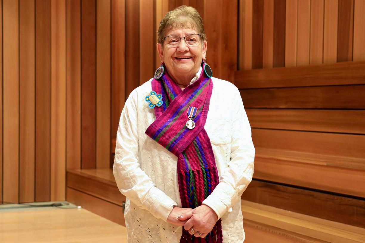 Métis Elder-in-Residence Stella Johnson standing in a cedar panelled room wearing a red and purple tartanMétis scarf with a King Charles III Coronation Medal