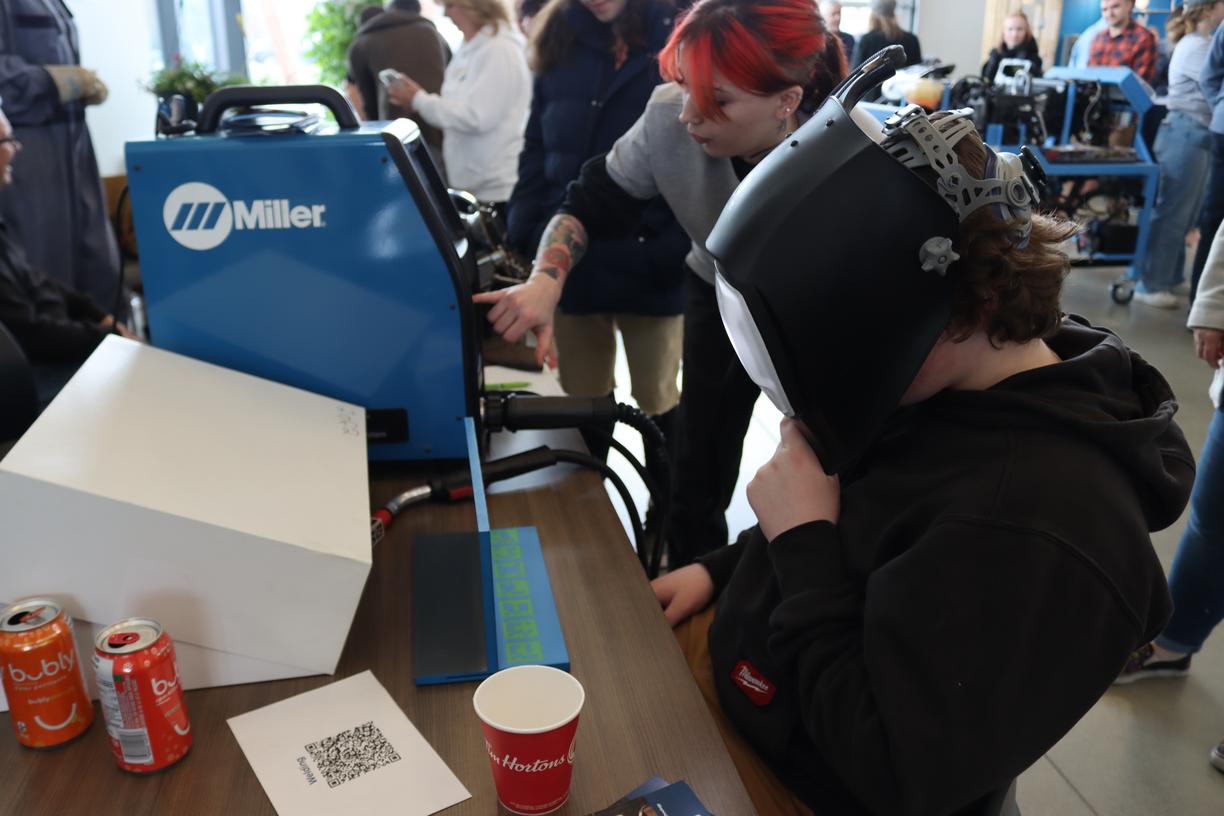 A person wearing a welding helmet is sitting at a desk trying a welding simulator machine