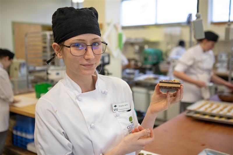 A person wearing a chef hat and chef jacket is holding up a Nanaimo bar dessert.