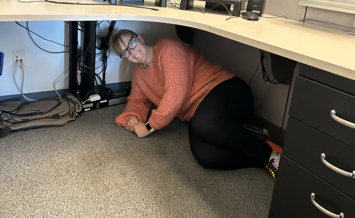 A VIU employee sheltering under a desk and smiling at the camera.