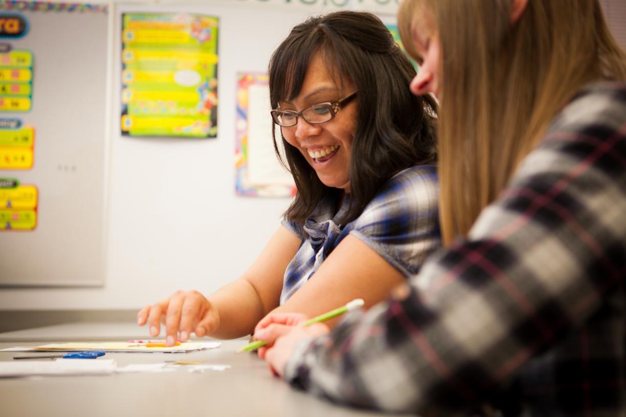 two students in a classroom looking at a sheet on a desk and smiling