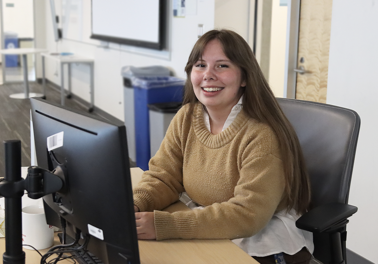 Kaylah Vassallo sittiing at a desk in front of a computer monitor and smiling at the screen.