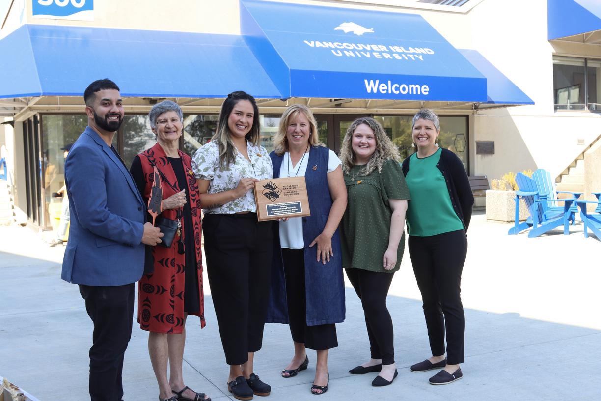 A group stands in front of the welcome centre holding a plaque