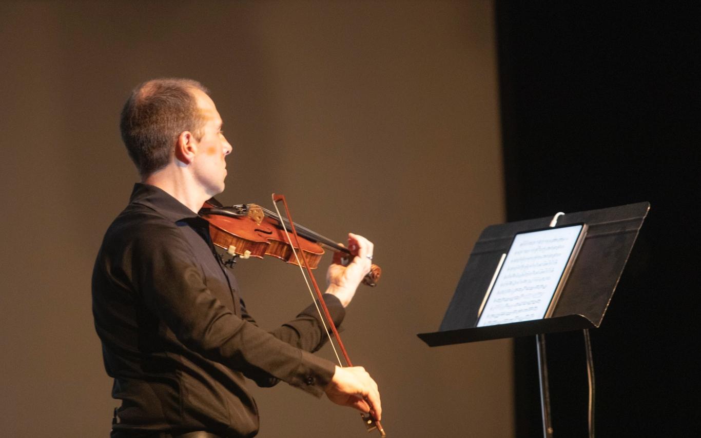 James Mark plays a violin while looking at a stand with sheet music on it.